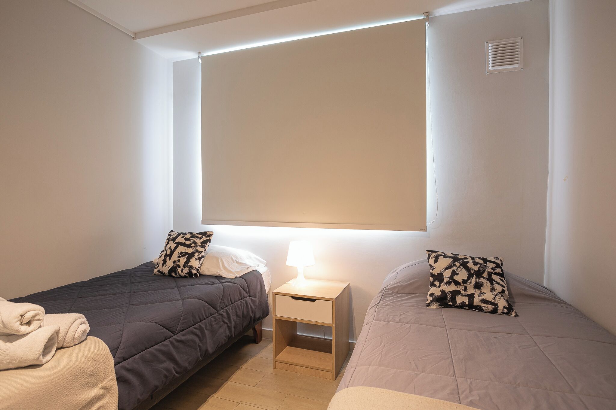 Une chambre avec deux lits simples, une table de chevet avec une lampe et une fenêtre avec des stores.