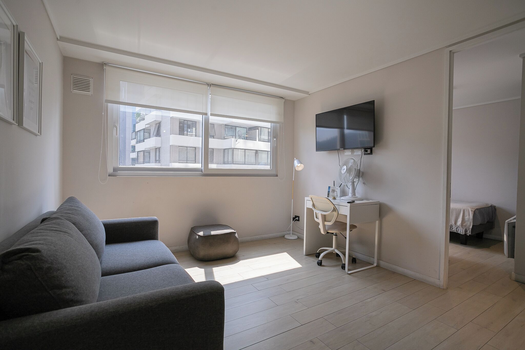 Un salon moderne avec un canapé gris, un petit pouf rond, un bureau avec une chaise, un téléviseur fixé au mur et une fenêtre avec des stores.