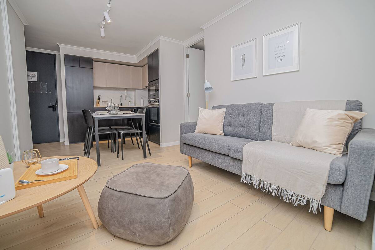 Un salon moderne avec un canapé gris, un petit pouf, une table à manger avec une tasse et une soucoupe blanches, et une cuisine en arrière-plan.