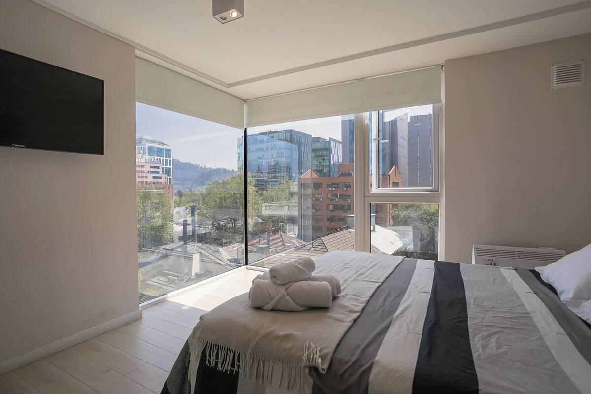 Une chambre moderne avec un grand lit, un téléviseur à écran plat fixé au mur et une vue sur la ville grâce à de grandes fenêtres du sol au plafond.