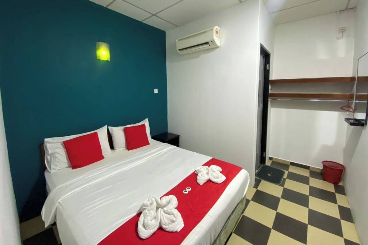Habitación de hotel con una cama, un juego de almohadas rojo y blanco, una toalla rojo y blanco, piso a cuadros, aire acondicionado montado en la pared y un estante con una cesta roja.