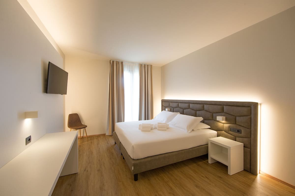 A modern hotel room with a large bed, bedside tables, a flat-screen TV mounted on the wall, and a wooden floor.