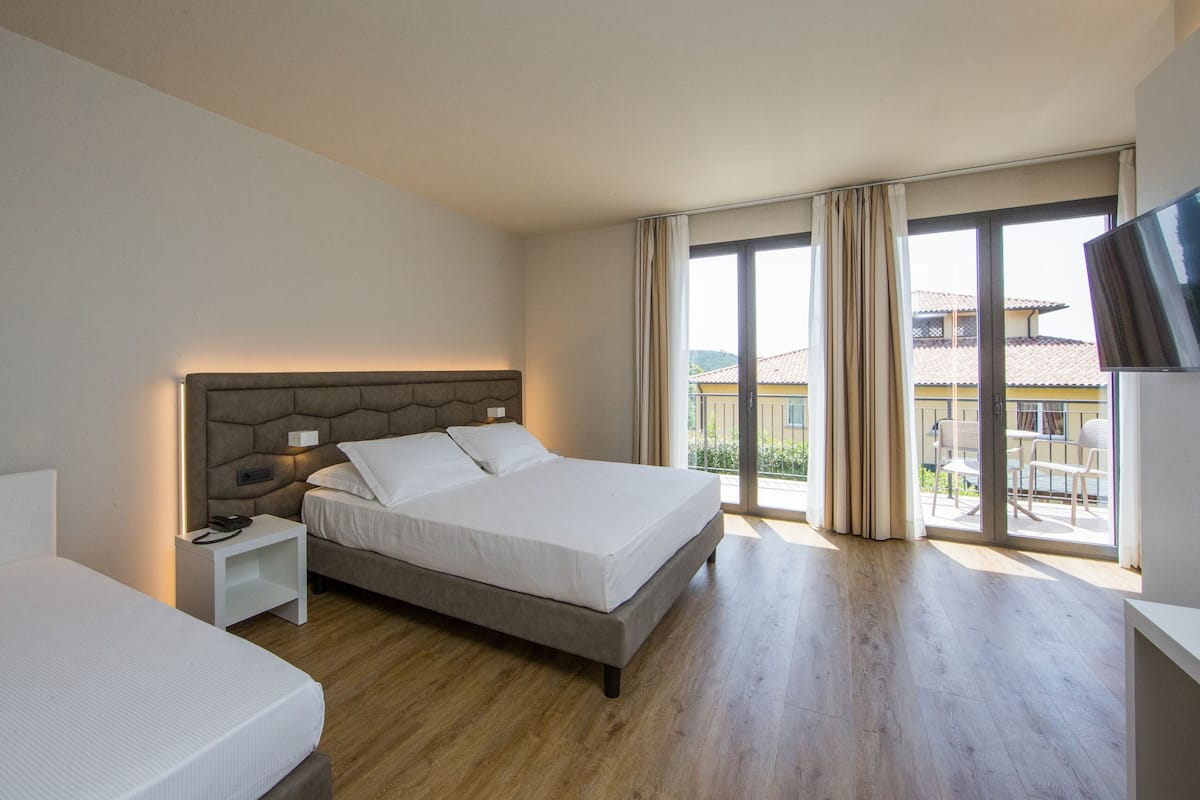 A hotel room with a large bed, a bedside table, a TV, and a balcony with a view of the outside.