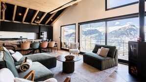 A modern living room with a high ceiling, large windows offering mountain views, a cozy seating area, and a fireplace. - Chalet, 100m from ski slopes, Ground floor, view mountain, sauna, terrace, balcony (La Plagne)