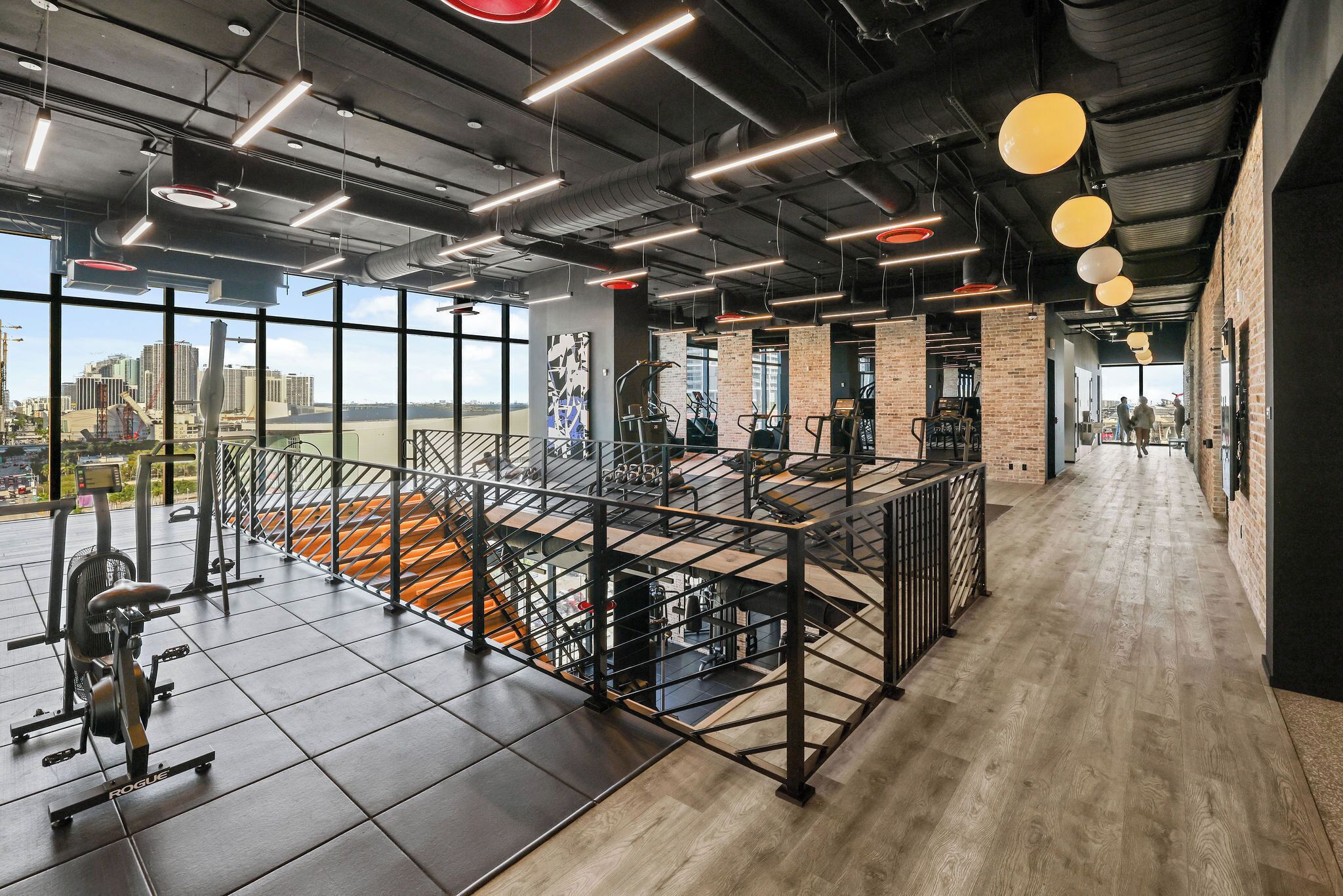 Un gimnasio espacioso con diversos equipos de ejercicio, amplias ventanas que ofrecen vistas de la ciudad y un techo alto con conductos expuestos.