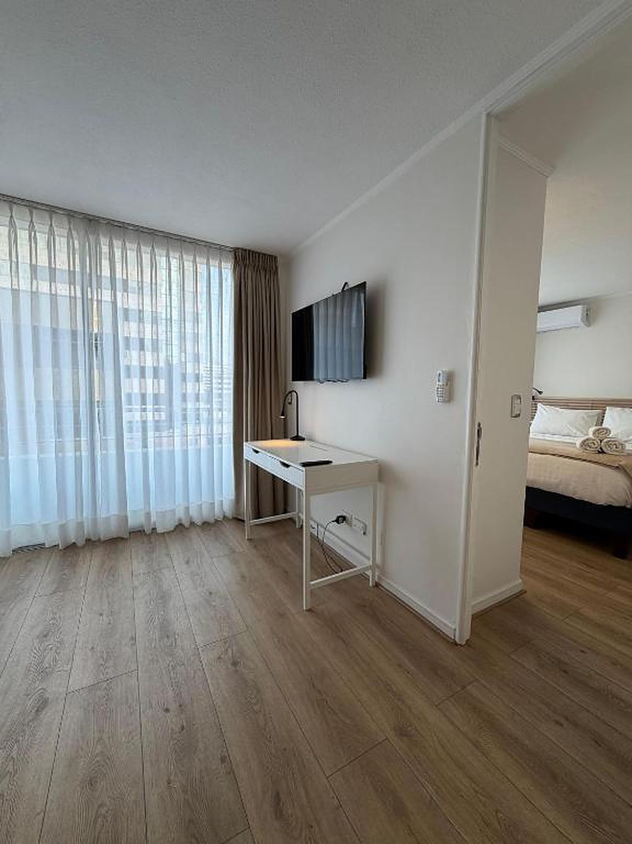 Una habitación de hotel con suelo de madera, un escritorio con silla, una cama y un televisor montado en la pared.