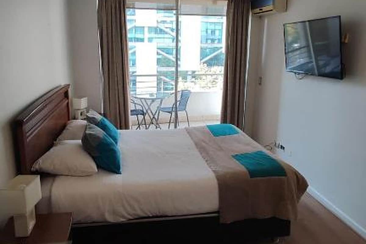 A hotel room with a bed, a TV, a balcony with chairs, and a view of buildings.