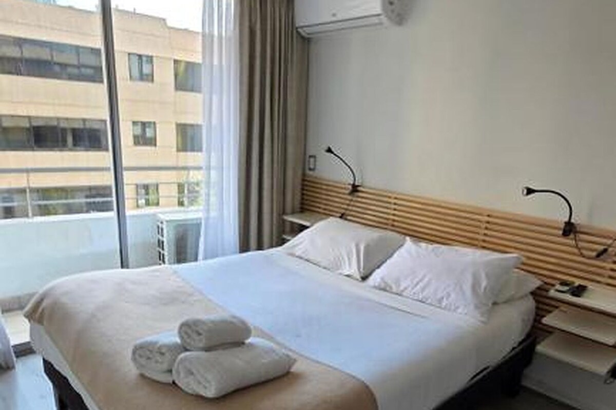 A hotel room with a bed, a bedside table, a lamp, and an air conditioning unit.
