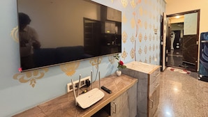 A modern living room with a large flat-screen TV mounted on the wall, a wooden console table with a router and a vase with a single red rose, and a patterned wallpaper. - Visla Stay - Premium Serviced Apartment  (Bengaluru)