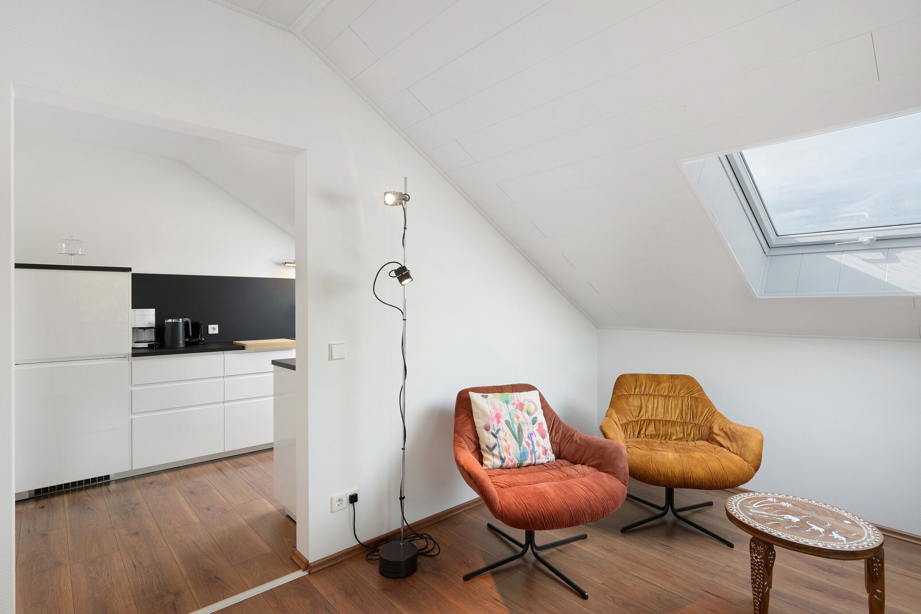 A modern kitchen with white cabinets, a black countertop, and a coffee machine. There are two chairs, one orange and one mustard-colored, with a small round table beside them. A floor lamp is visible, and the room has a sloped ceiling with a skylight.