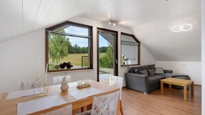 A modern loft with a dining area, a gray sofa, and large windows overlooking a green landscape.