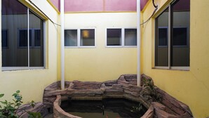 Un pequeño estanque con rocas y una estructura de madera, rodeado por un muro amarillo con ventanas.