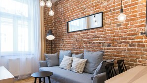 A cozy room with a brick wall, a grey sofa with patterned cushions, a small round table, and a wooden dining table. - 19Rooms (Kraków)