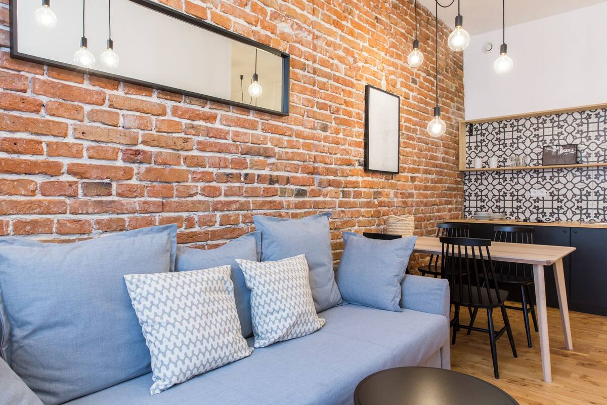 Un espacio moderno de estar con una pared de ladrillo, un sofá con cojines estampados, una zona de comedor con una mesa y sillas, e iluminación colgante.