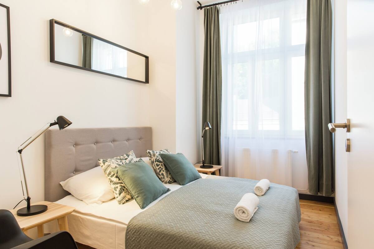 Habitación de hotel ordenada con cama, mesita de noche, lámpara y un amplio ventanal con cortinas.
