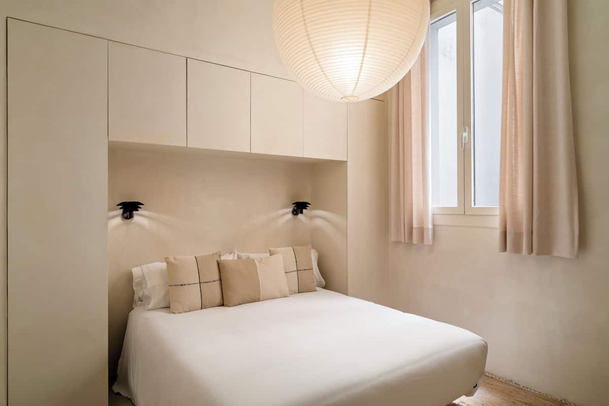 A bedroom with a bed, headboard, wall-mounted light, and window with curtains.