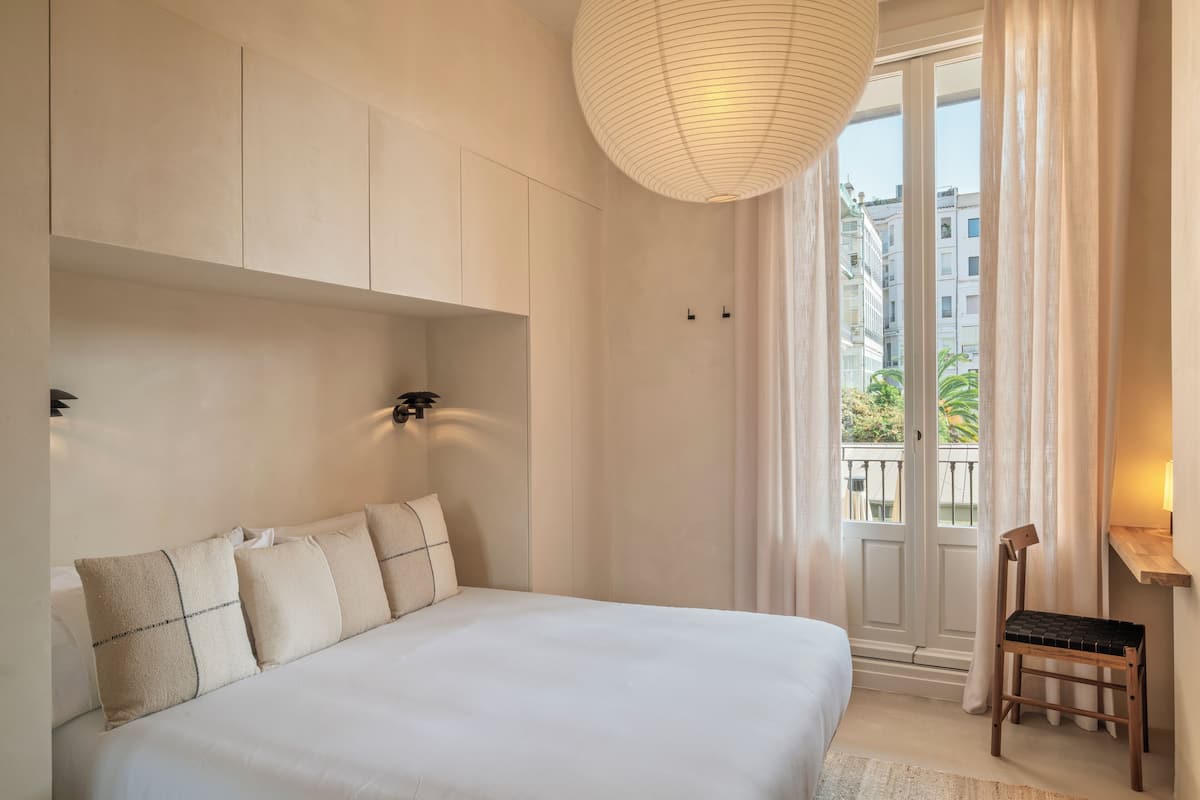 A bedroom with a large bed, a wooden chair, a hanging light fixture, and a view of buildings through a door.