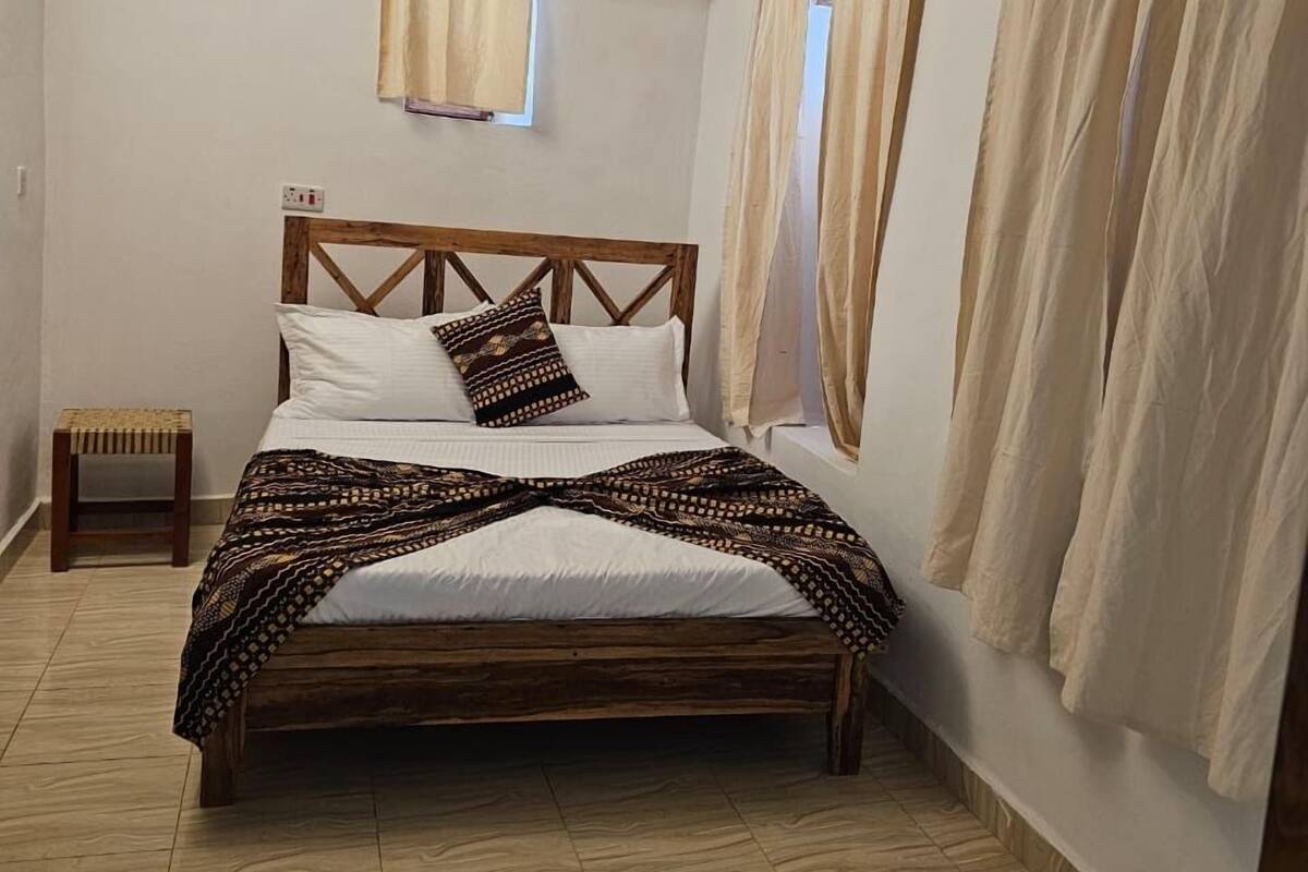 Un dormitorio con cama de madera, cortinas color arena, una mesita de noche pequeña y un tapete tejido.
