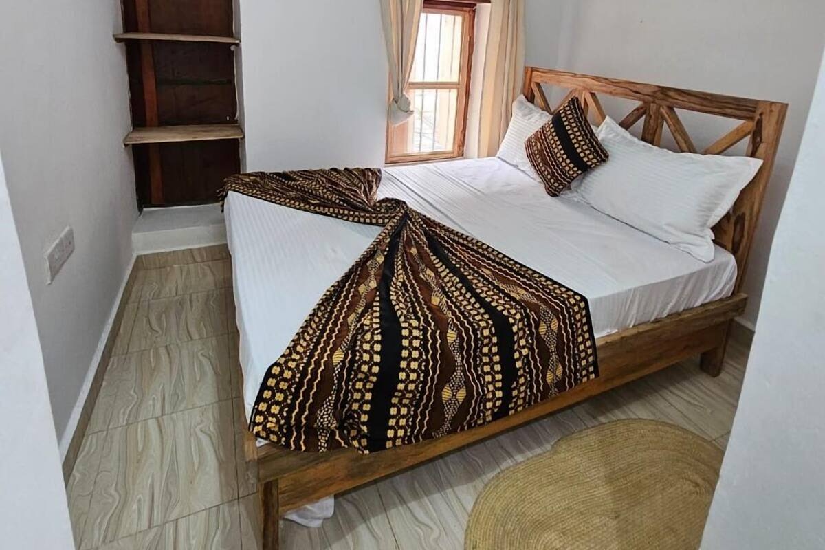 Un dormitorio con cama de madera, ropa de cama blanca, ventilador de techo, ventana con cortinas y una pequeña estantería.