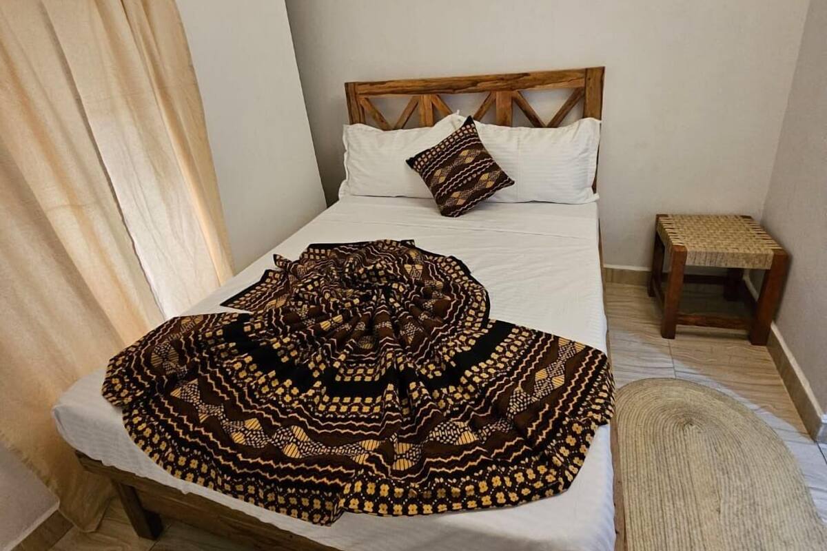 Un dormitorio con estructura de cama de madera, ropa de cama blanca, una manta estampada, un taburete de madera pequeño y una alfombra tejida.