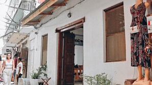 A narrow street with a white building featuring a wooden door and windows. A mannequin dressed in a patterned dress is displayed outside.