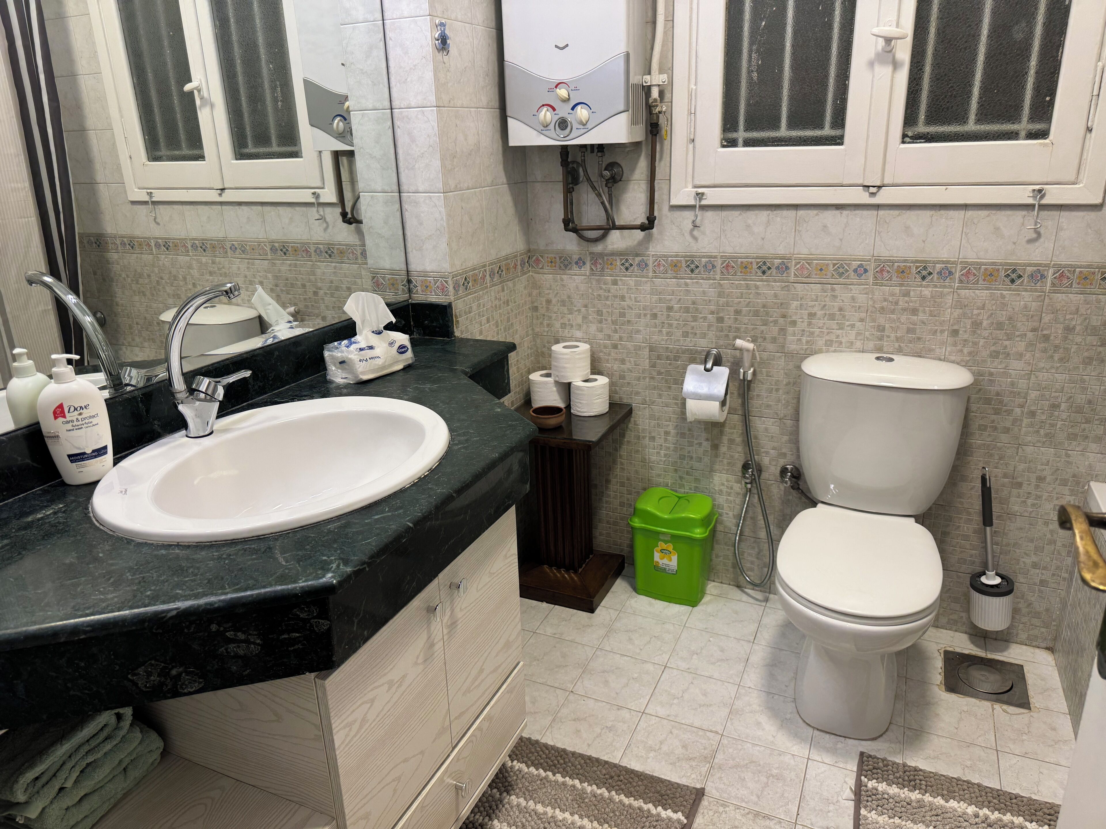 A bathroom with a sink, toilet, and shower. There are toiletries on the counter and a green trash bin.