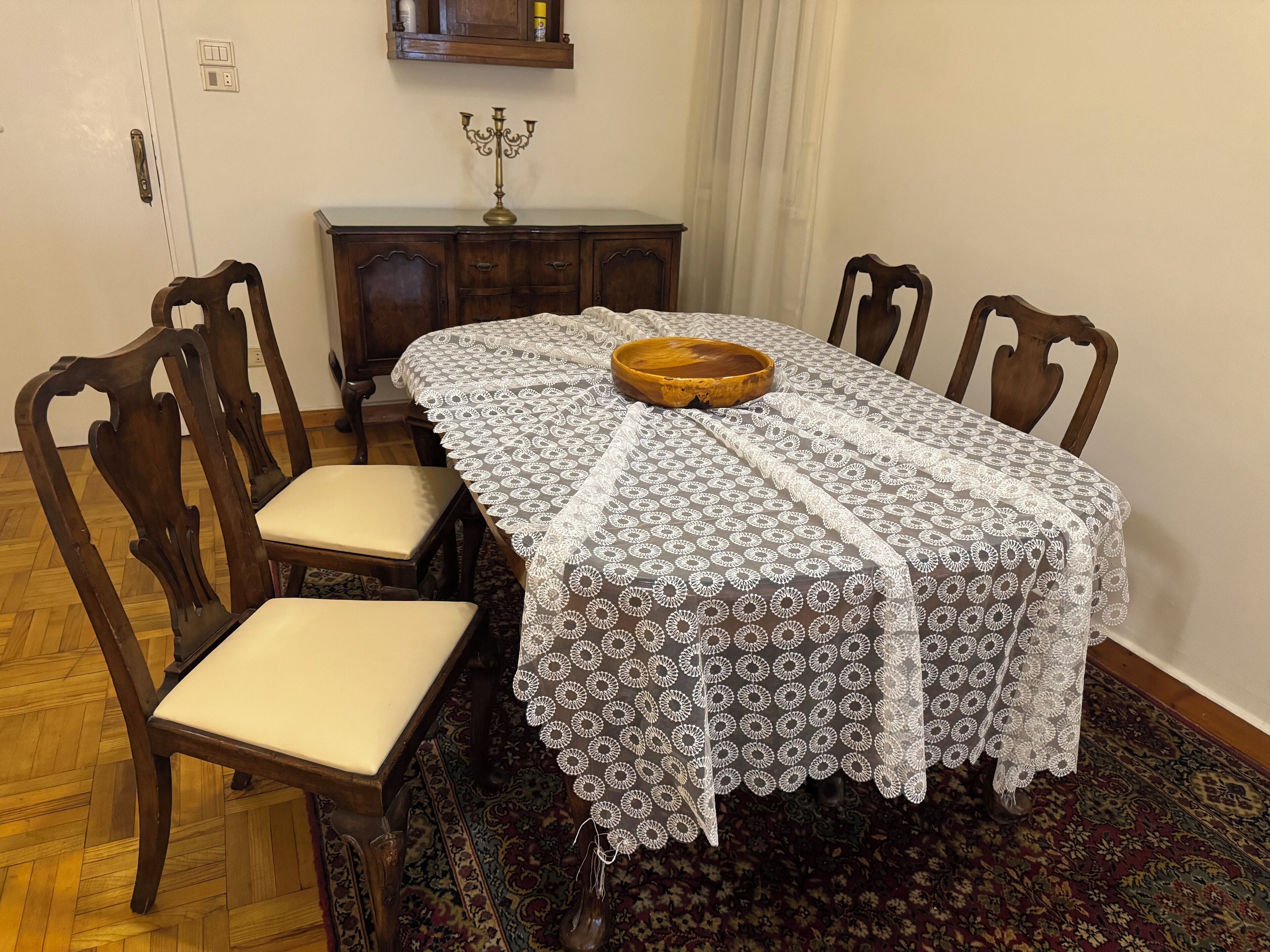 A dining area with a table covered in a lace tablecloth, surrounded by wooden chairs with cushioned seats.