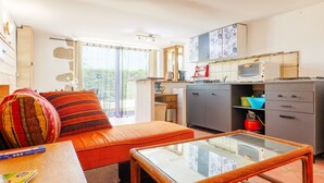 Un espace de vie compact comprenant un coin cuisine, une table à plateau en verre, un canapé avec des coussins et une vue sur la verdure par les fenêtres.