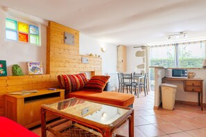 A compact living space with a sofa, a glass-top coffee table, a dining area with chairs, a kitchen with a microwave, and a small cabinet.