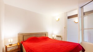 Un lit simple avec une couverture rouge, une table de chevet avec une lampe et une fenêtre avec des stores.