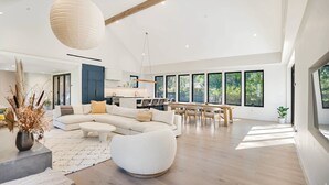 Un'ampia zona living con un grande divano a forma di L, un poggiapiedi rotondo e un tavolo da pranzo con sedie. È presente un angolo cottura con un pensile blu e un lampadario.