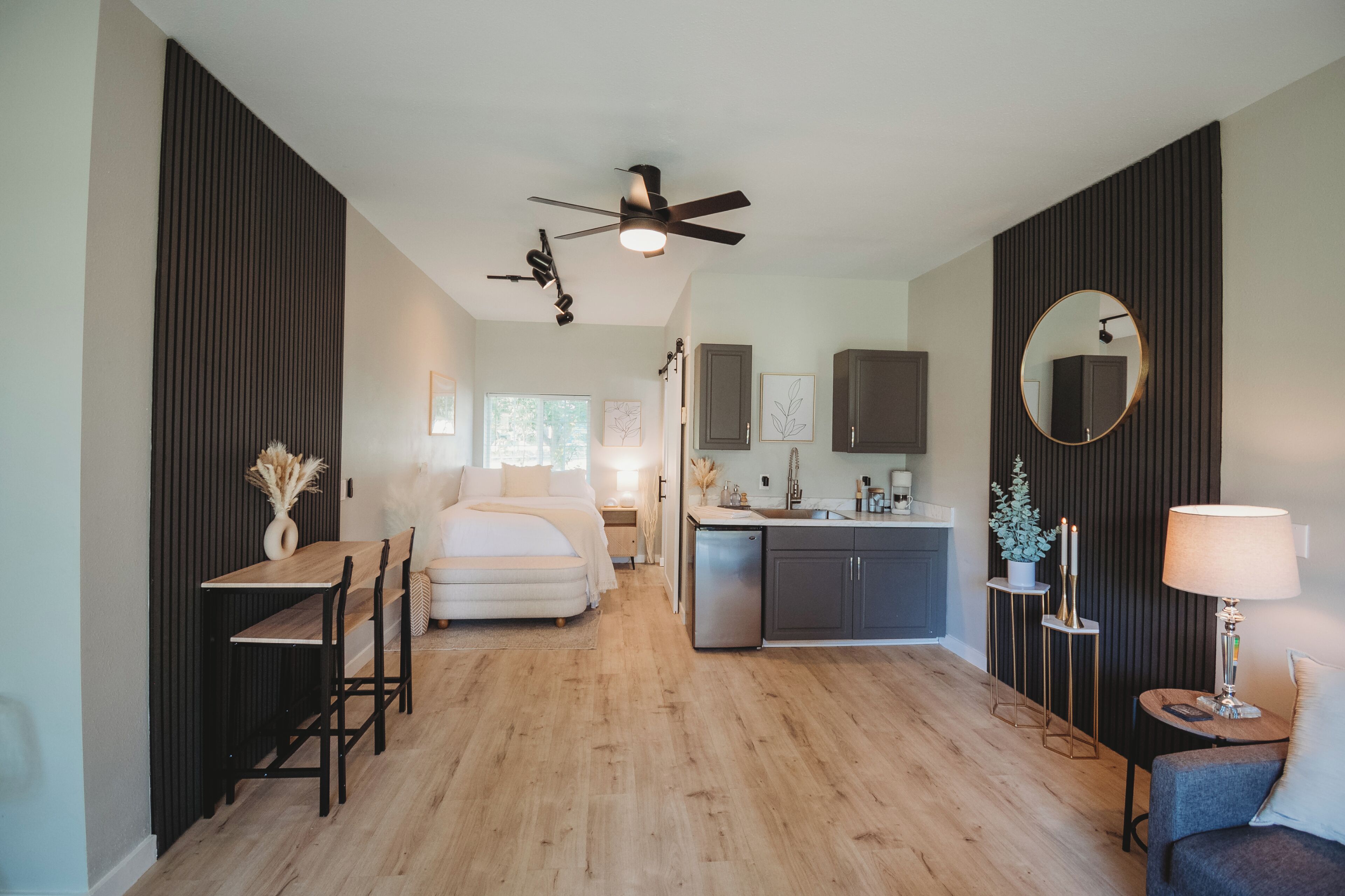 Una habitación de hotel moderna con suelo de madera, una cama, un escritorio con silla, una pequeña cocina y un ventilador de techo.