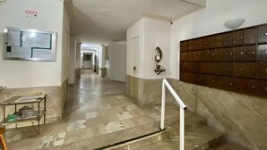 Un pasillo con piso de mármol, barandal blanco, un tablón de anuncios y una caja de seguridad en la pared.