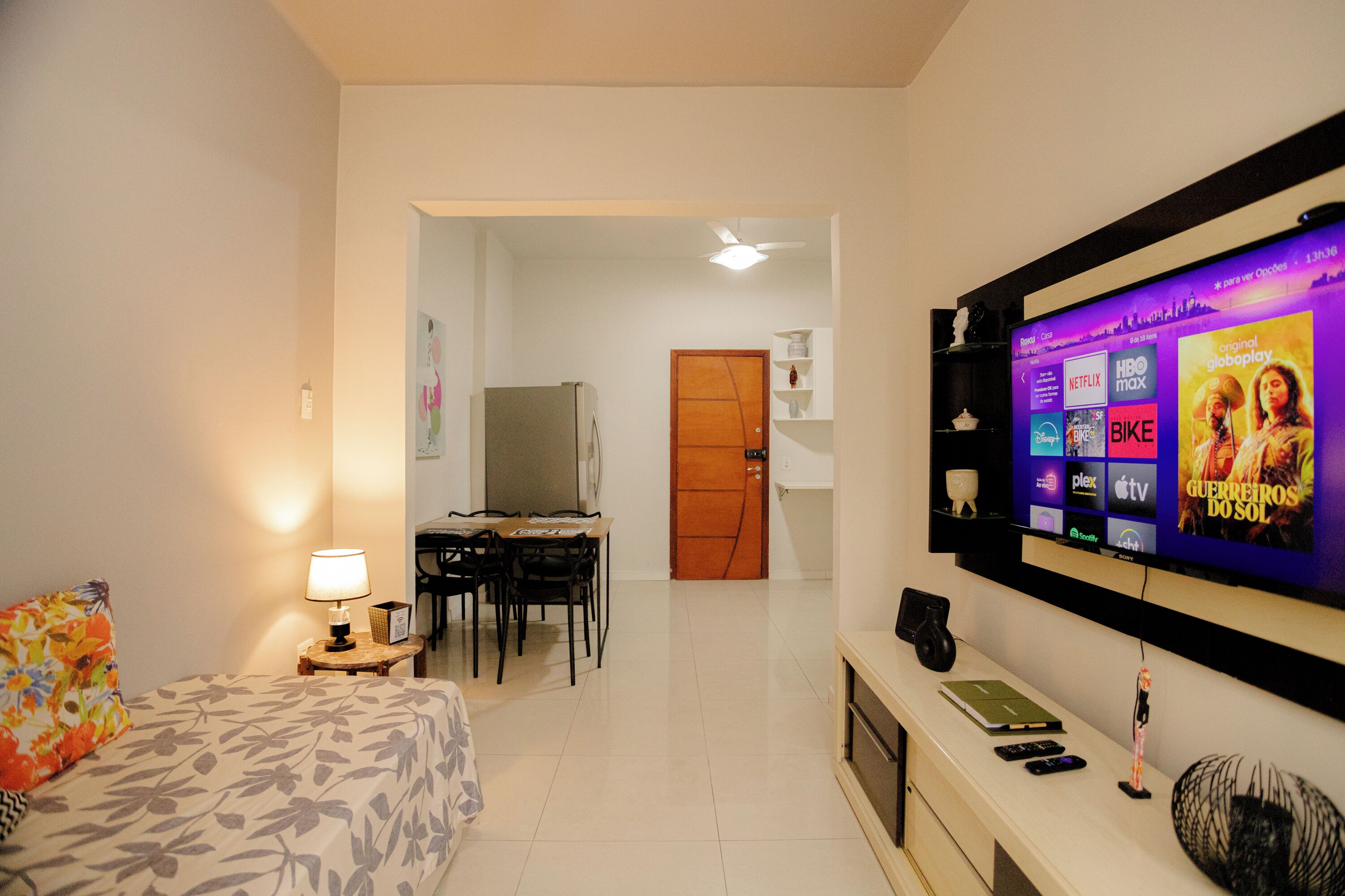 A hotel room with a bed, a desk, a television displaying a movie, and a small table with a lamp.