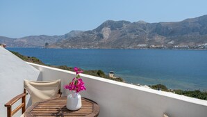 Ein Balkon mit einem Holztisch und einer weißen Vase mit rosa Blumen, mit Blick auf eine Küstenlandschaft mit Bergen und dem Meer.