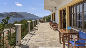 A terrace with tables and chairs, a view of the sea, and a mountainous landscape in the background.