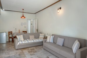 A living room with a sofa, a dining table, chairs, a refrigerator, and a hanging light fixture.