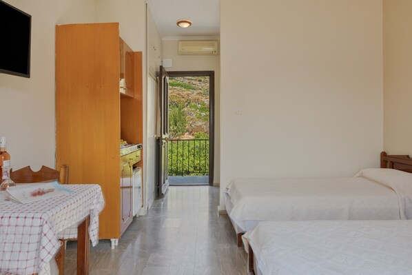 Une chambre d’hôtel avec un lit, une table recouverte d’une nappe à carreaux, un meuble en bois et une vue sur un balcon agrémenté de plantes.
