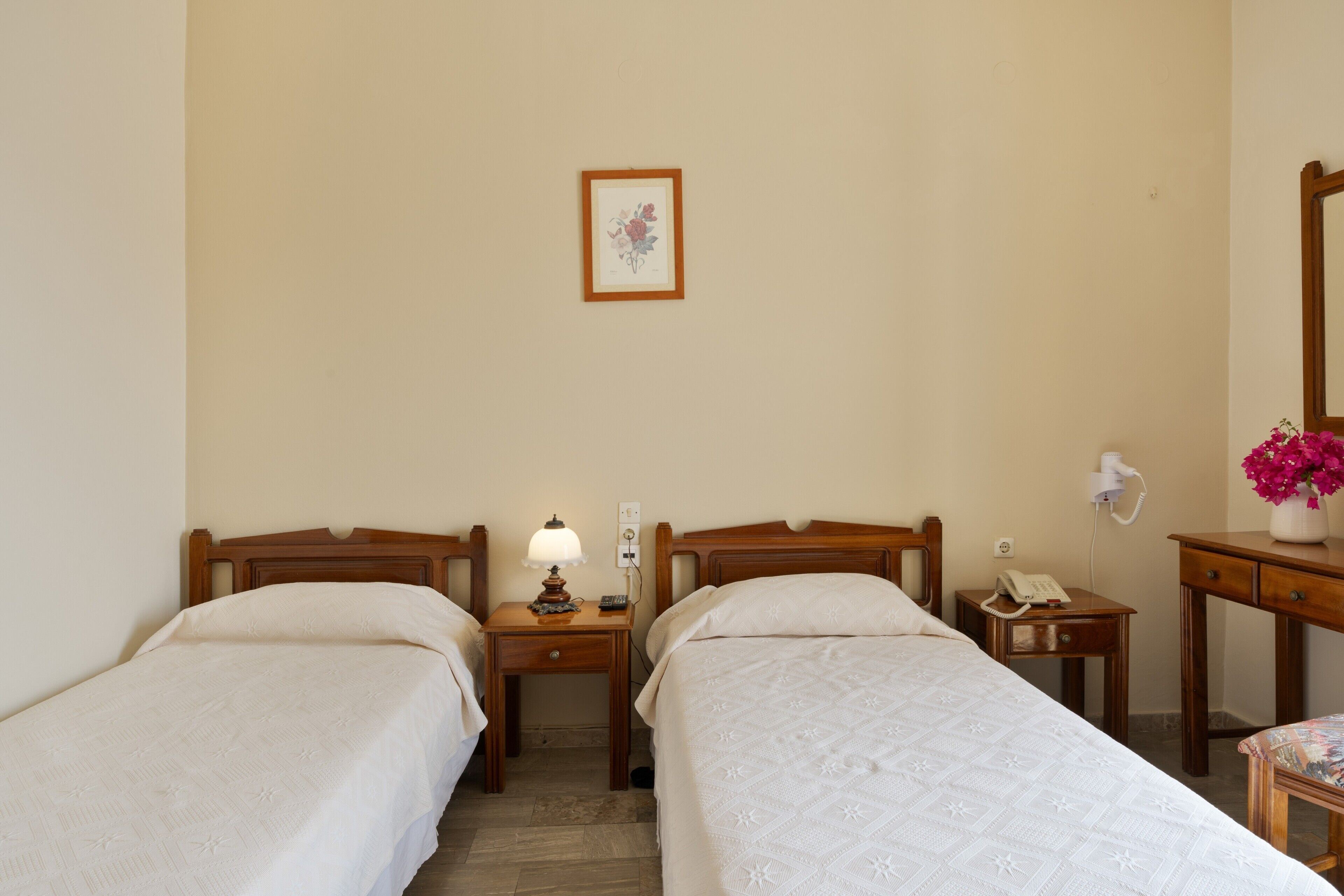 A hotel room with two single beds, a bedside table with a lamp, a telephone, a small table with a vase of flowers, and a wall-mounted picture.