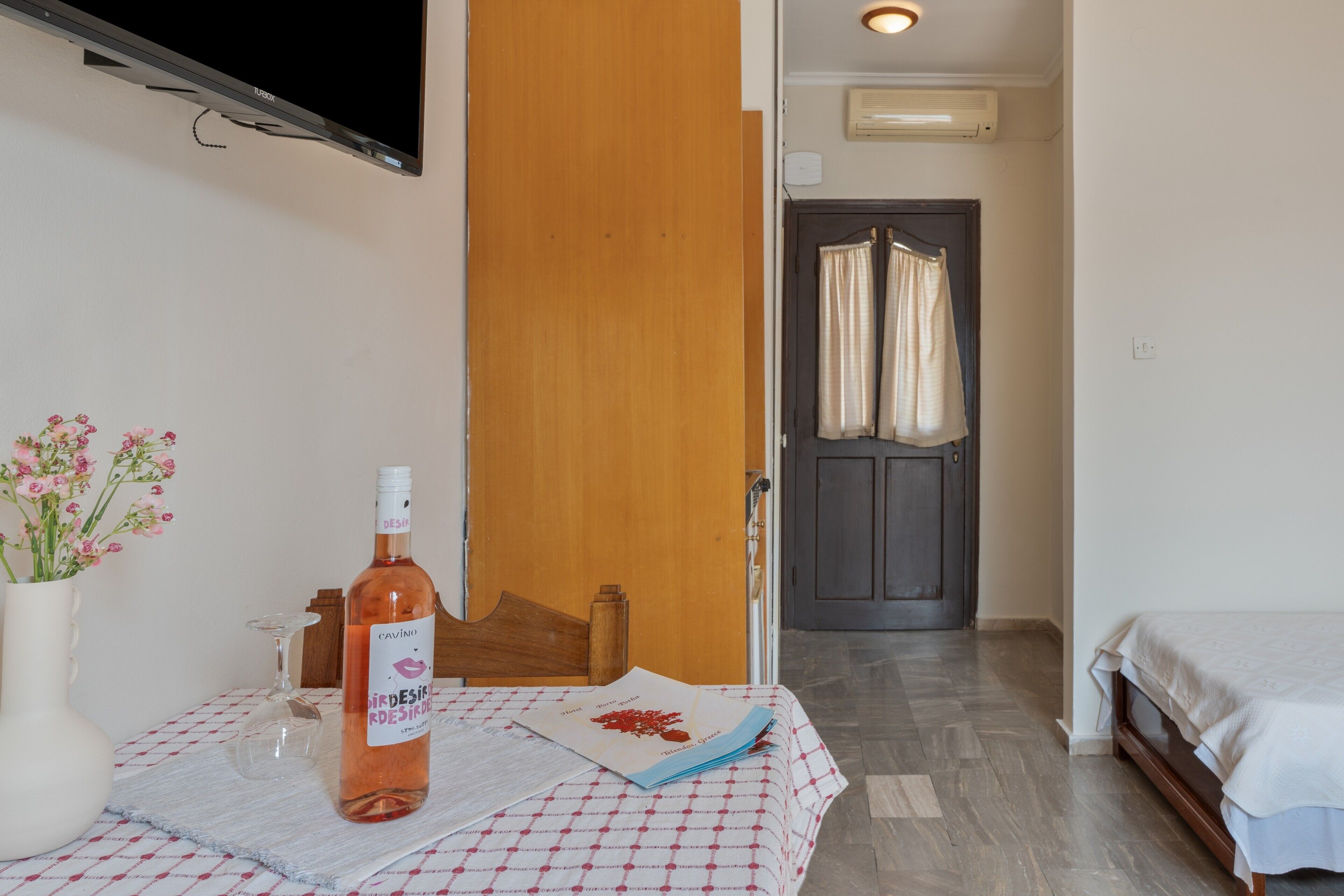 A hotel room with a bed, a table with a bottle, a vase with flowers, and a door leading to another room.