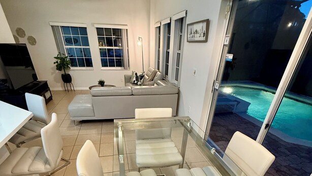 Un espace repas moderne avec une table à plateau de verre et des chaises blanches, un canapé gris et une vue sur une piscine la nuit.