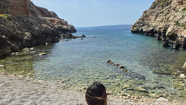 Une personne assise sur une plage rocheuse, avec une eau claire et des falaises en arrière-plan.