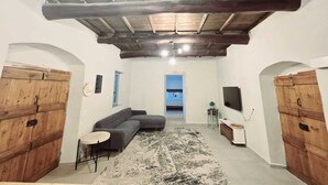 Une pièce avec un plafond en bois, un canapé gris, une petite table ronde et un téléviseur posé sur un meuble.