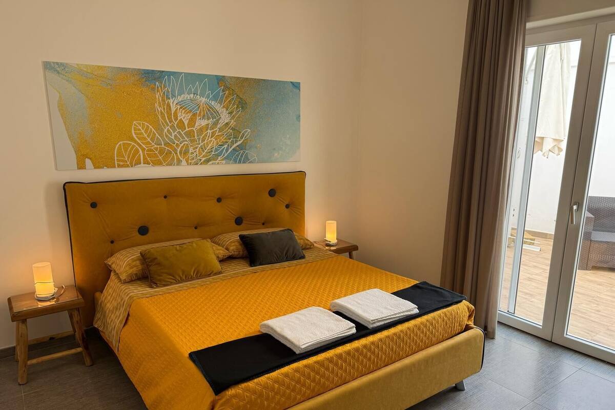 Un cuarto de hotel con una cama grande, dos mesitas de noche, una lámpara de noche, un ventanal con cortinas y un cuadro en la pared.