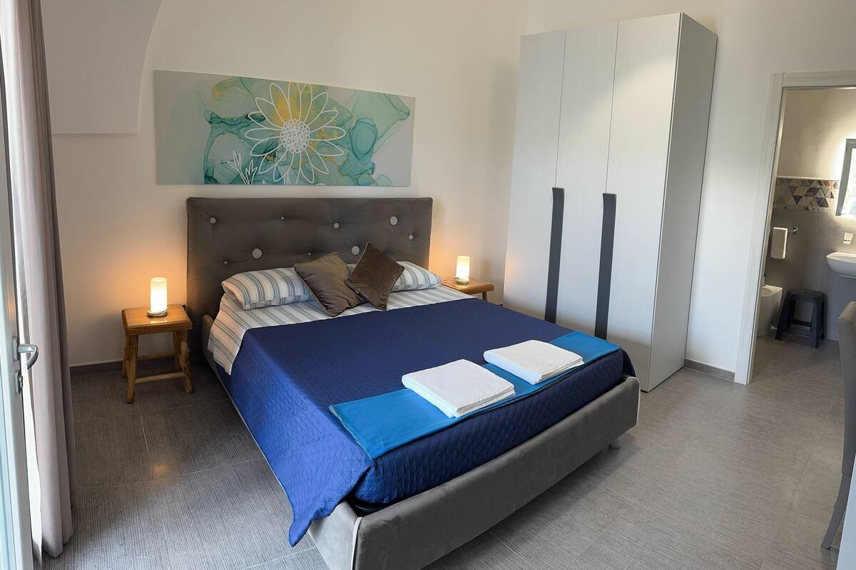 Un dormitorio con una cama grande, cabecera, mesita de noche, lámpara de noche, armario y un baño visible a través de una puerta abierta.