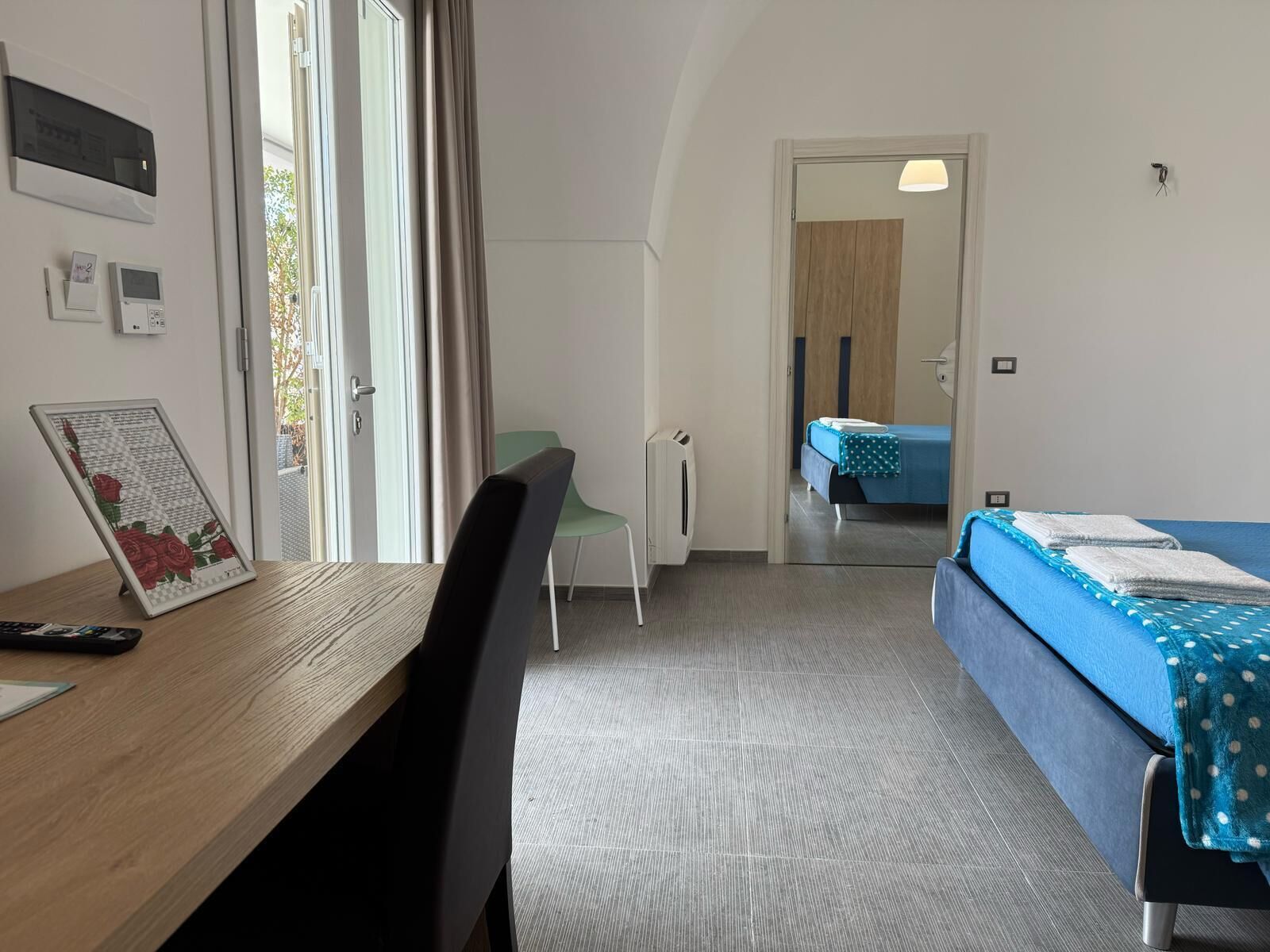 Una habitación de hotel moderna con un escritorio de madera, una cama individual con ropa de cama azul, una silla verde y una mesita de noche pequeña.