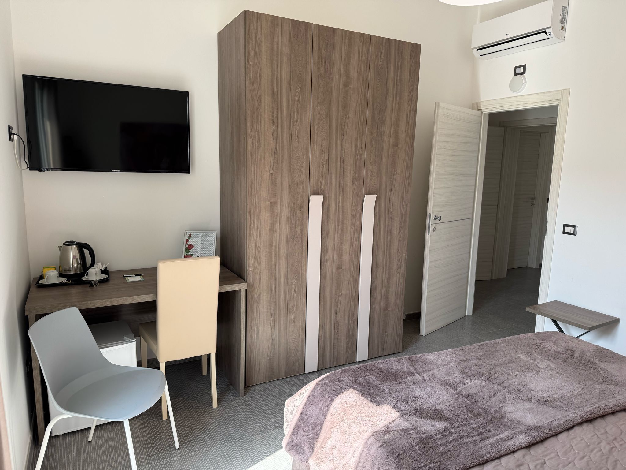 A modern hotel room with a wooden wardrobe, a desk with a kettle and cups, a grey chair, and a bed with a blanket.