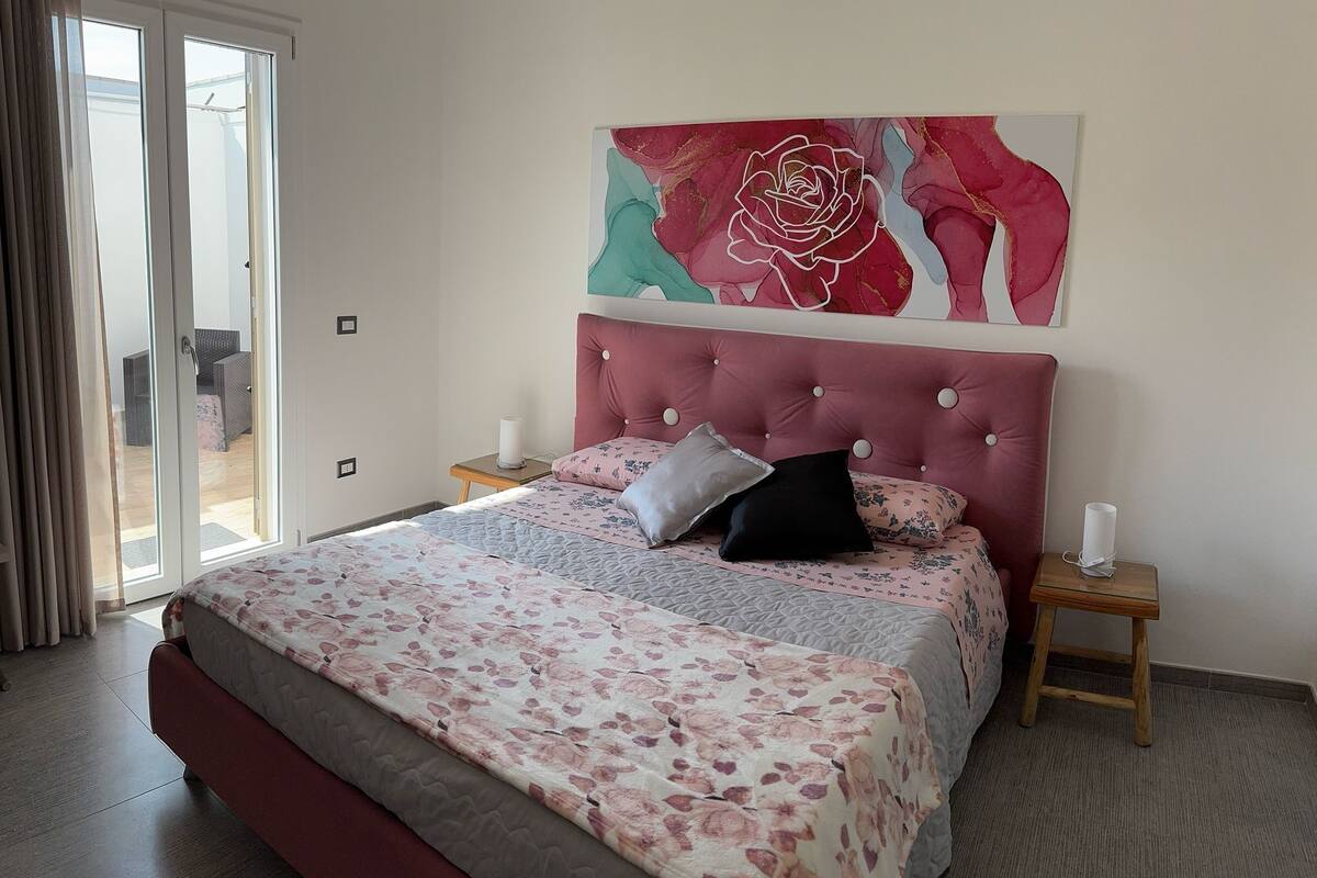 Un dormitorio con una cama grande, cabecera roja y colcha con estampado floral. Encima de la cama hay un cuadro abstracto de gran tamaño y, a la izquierda, una ventana con cortinas.