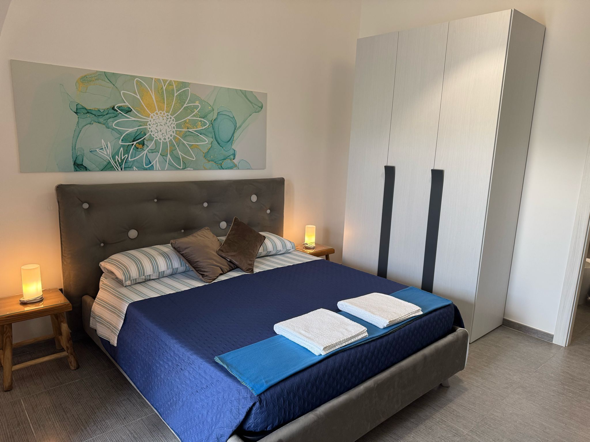 A bedroom with a bed, a nightstand, a bedside lamp, a wall art piece, and a wardrobe.