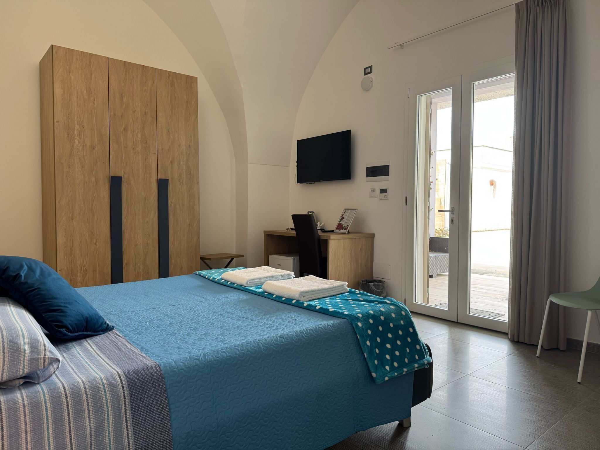 Une chambre avec un lit, un bureau, une t élévision et une porte-fenêtre coulissante donnant sur un balcon.